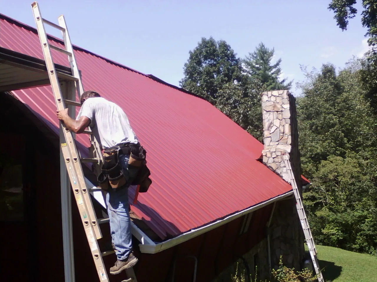 Roofer on a ladder beside a red metal roof and stone chimney — Soffit & Fascia Repair by Roofing Brothers in Accident